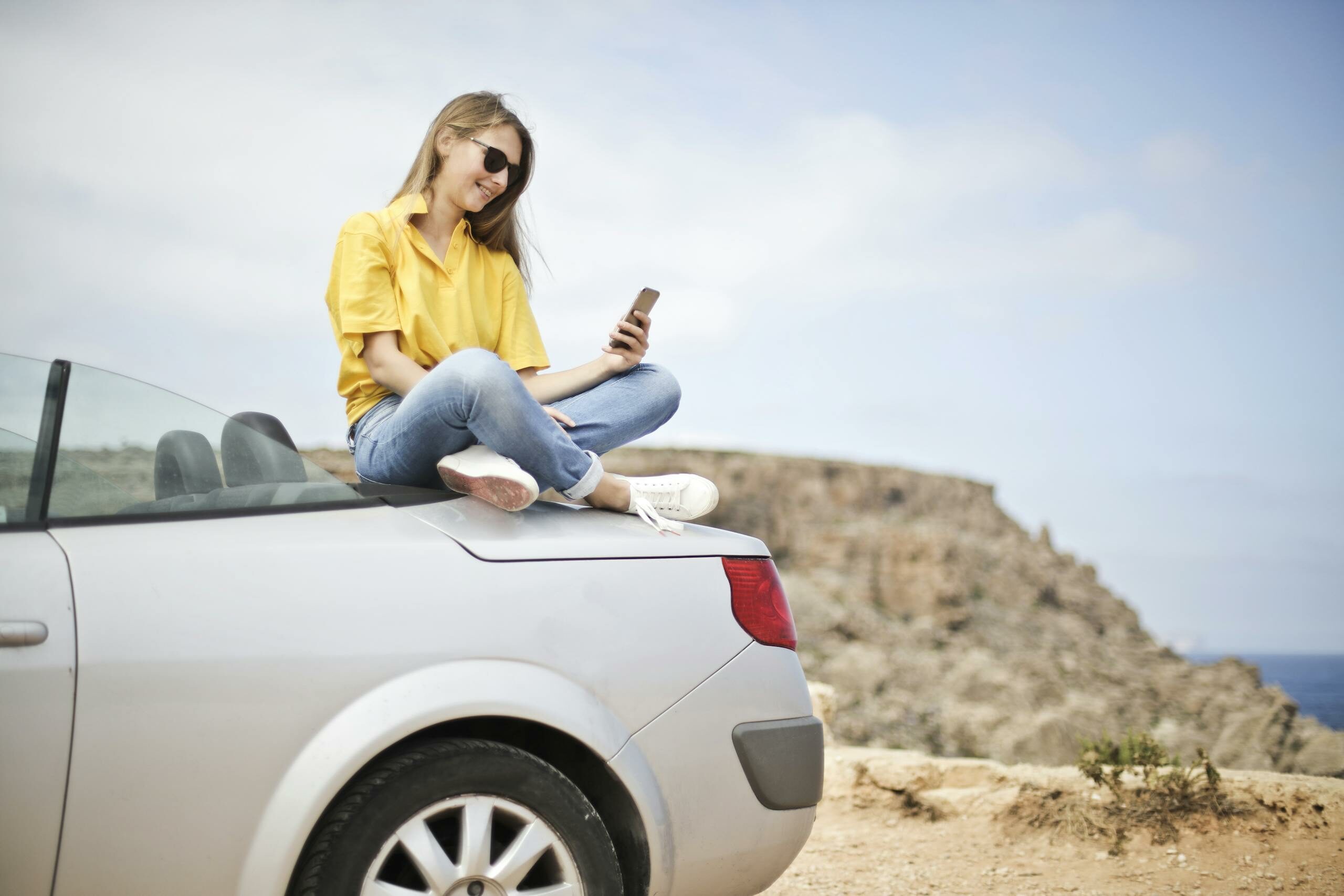 Woman in Yellow Blouse and Blue Jeans Taking Selfie While Sitting on Car